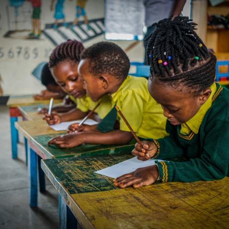 A little girl, Freza 5, in a classroom in Rwanda sitting at a desk, other children in the background