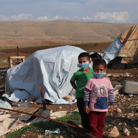 A little boy and a little girl wearing facemasks stand in front of tents destroyed by Israeli forces in Palestine